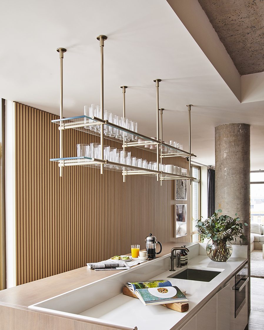 Modern, bright and earth toned kitchen with 1 ceiling mounted shelving unit in solid warm brass with champagne finish, 2 bay, hanging above kitchen island with sink, holding crystal glassware on 2 glass shelves.
