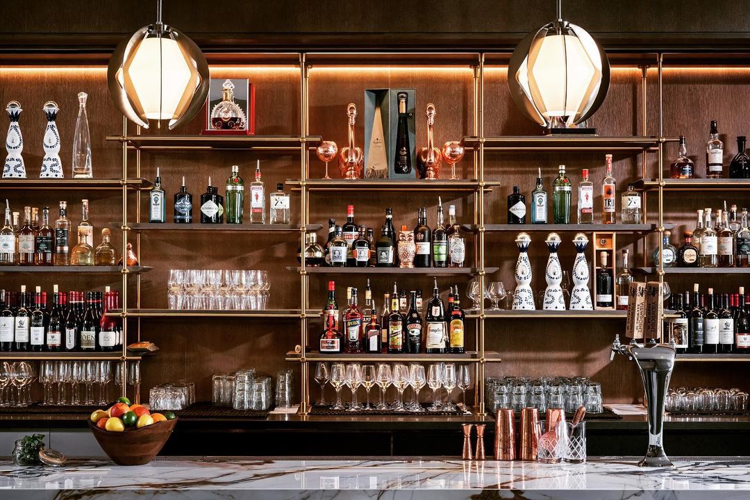 Dark, woody, cocktail bar showing ceiling mounted solid warm brass shelving unit and cerused oak shelves mounted to bar counter by Amuneal. Back bar features liquor display and glassware storage behind bar.