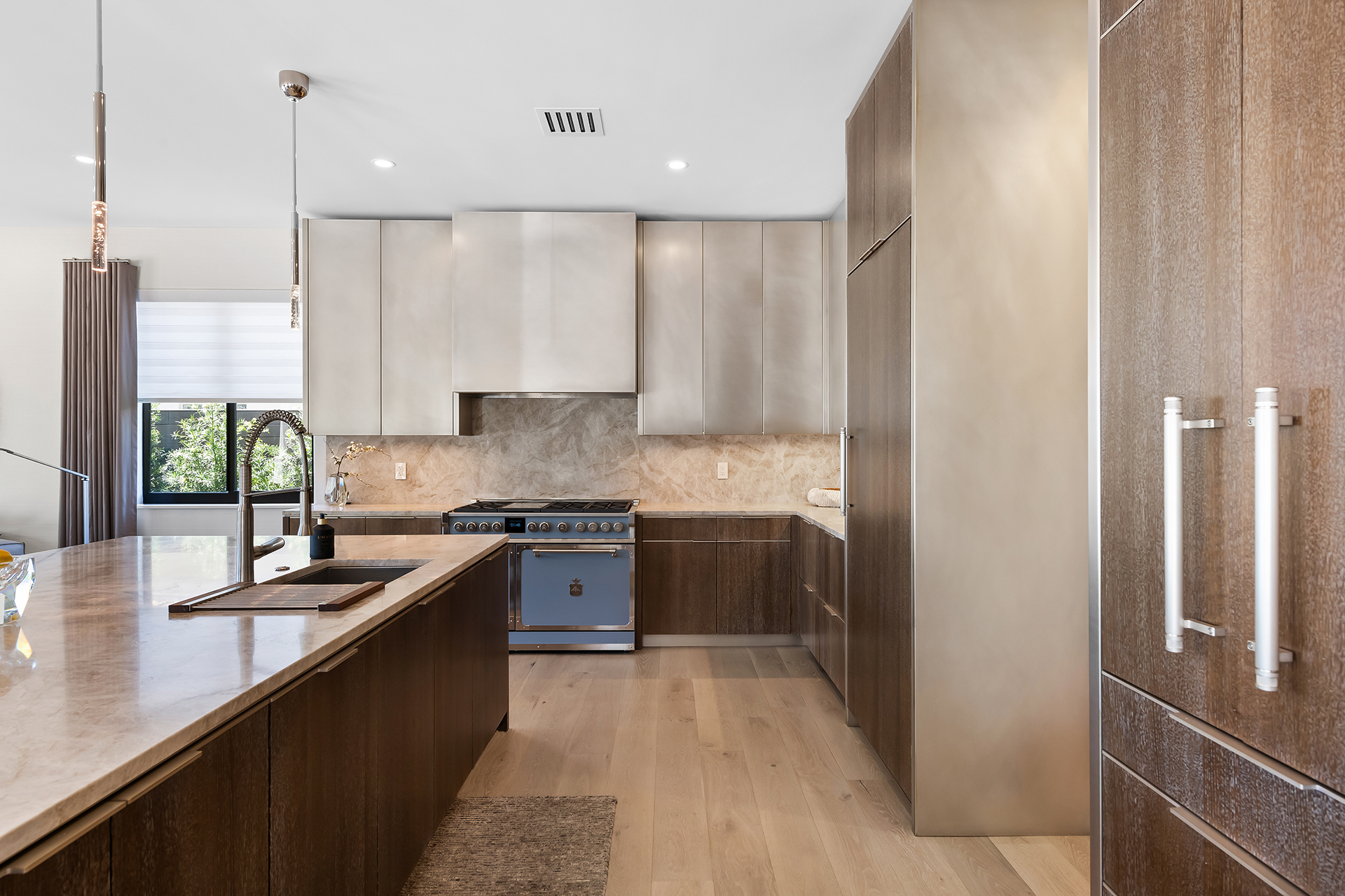 Full custom kitchen made by Amuneal with champagne brass upper cabinets, brass cladded range hood, cabinet doors and surrounds. An enclosed refrigerator, hidden appliances with large vertical metal door handles behind chestnut. Quartz kitchen island with sink on chestnut wood lower cabinets with champagne brass pulls horizontally along each door, matching metal kicks flush against a light tan, hardwood floor